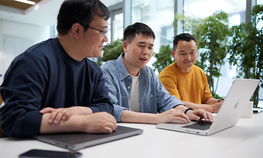 Ken 在 MacBook 上打字，兩邊各坐著一位同事，背景有幾扇光亮的窗戶和一些植物。