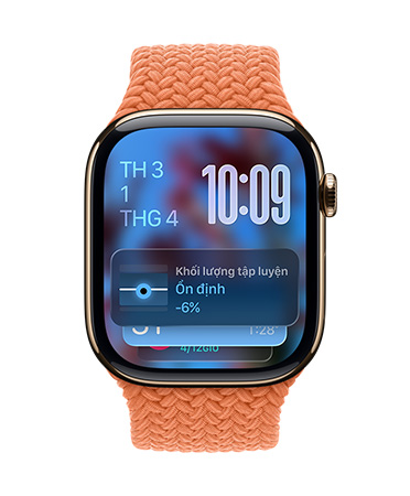 Apple Watch màu Vàng Hồng với dây bện màu Quýt Chín, mặt đồng hồ hiển thị ngày ở trên cùng bên trái, thời gian với các chữ số xếp chồng lên nhau ở trên cùng bên phải, tiện ích Khối Lượng Tập Luyện ở dưới cùng, tất cả đều được hiển thị dạng Liquid Glass trong mờ