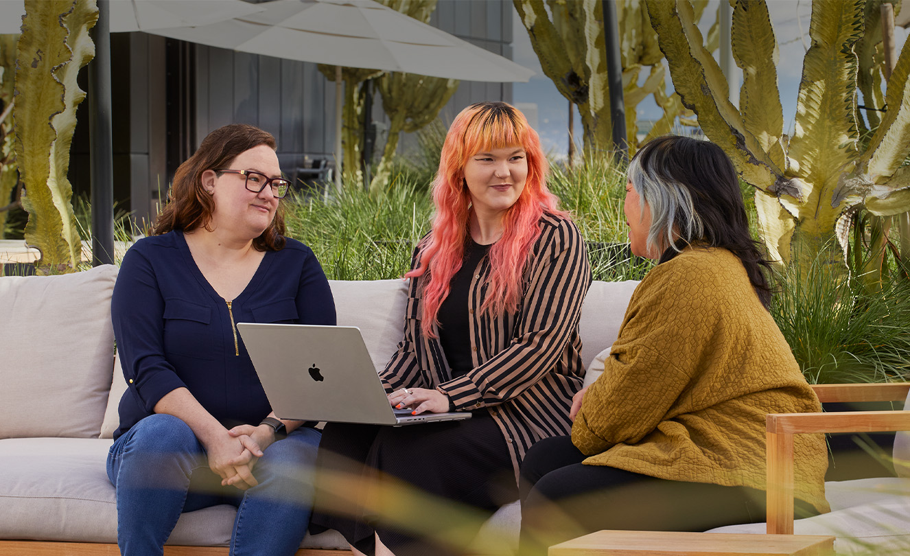 Tre Apple-ansatte sitter sammen utendørs på en terrasse. En av dem har en MacBook.