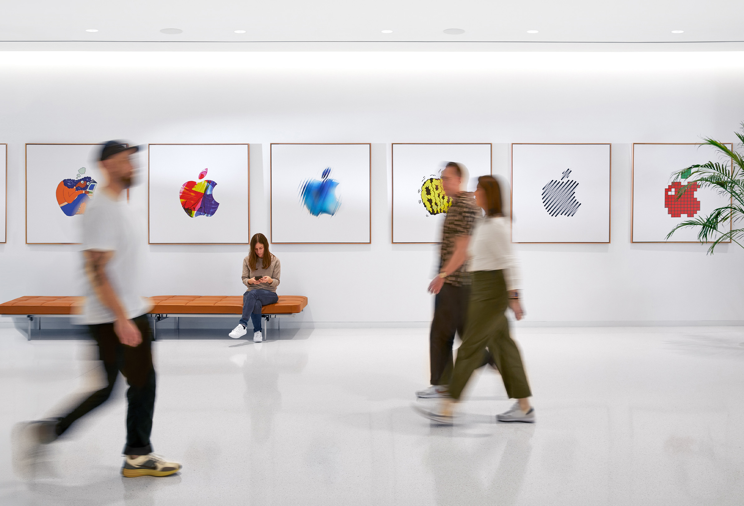 Tre Apple-medarbeidere går forbi en innendørs vegg med fargerike Apple-logoer, mens en fjerde ansatt sitter på en benk.