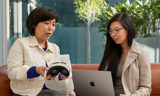 Jessica sitter innendørs og jobber på en MacBook med døren åpen ut til terrassen. En Apple-kollega viser henne Apple Vision Pro.