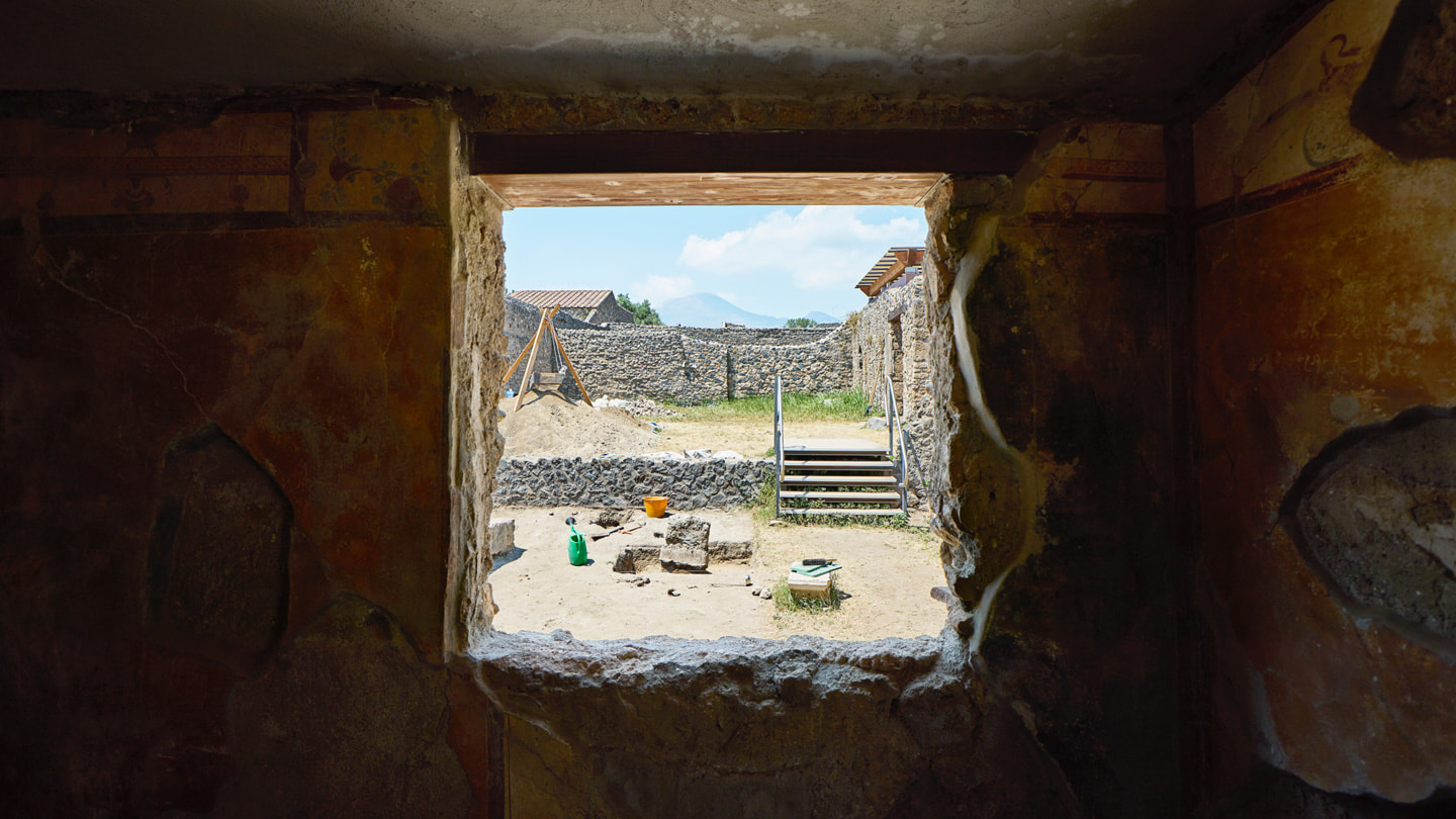 Vista a través de una ventana al sitio de la excavación muestra muros de piedra y el Vesubio a lo lejos. 