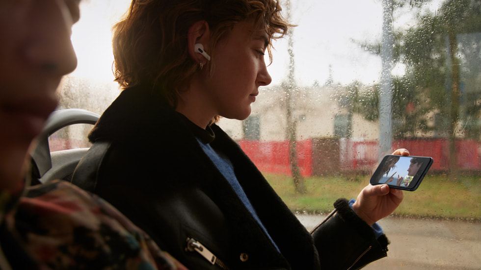 バスに乗っている少女がAirPodsを装着し、iPhone SEで、5Gを使用してSharePlayでビデオを観ている様子。